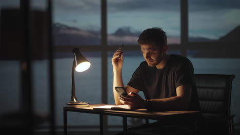 A-thoughtful-man-sitting-at-a-table-with-a-desk-lamp-and-a-mobile-phone.-The-dark-day-is-twilight-against-the-background-of-the-window-a-sad-man-is-thinking-about-work-while-sitting-at-home.